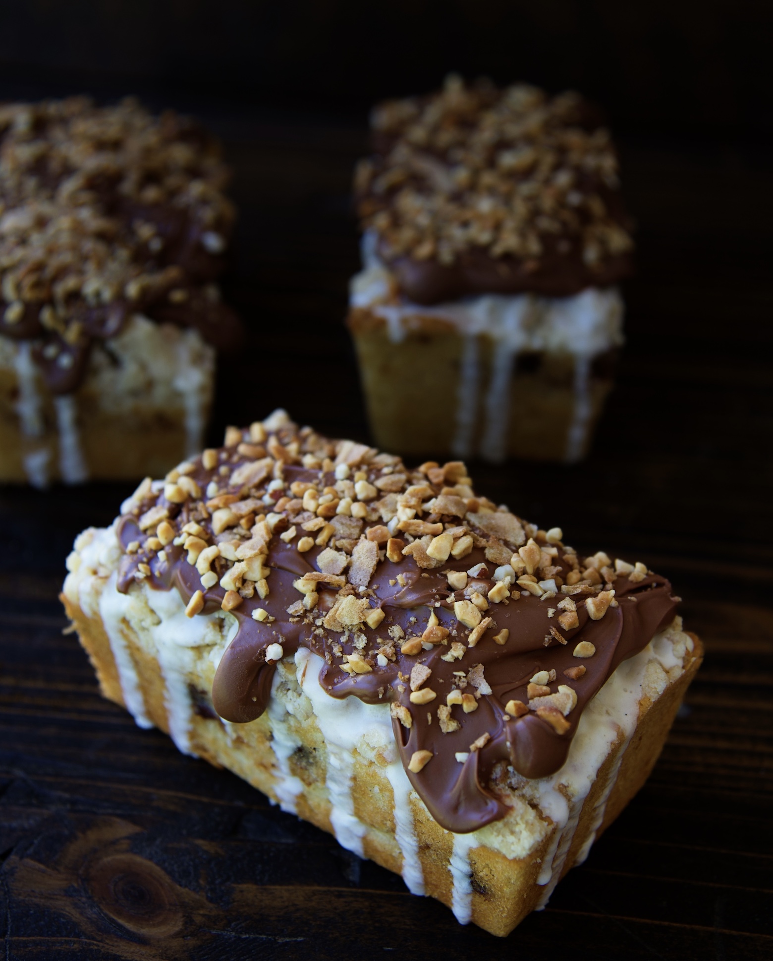 Three Glazed Drumstick Crumb Cakes with front one in focus. Covered in vanilla ice cream glaze and a chocolate coating topped with peanuts and sugar cone. 