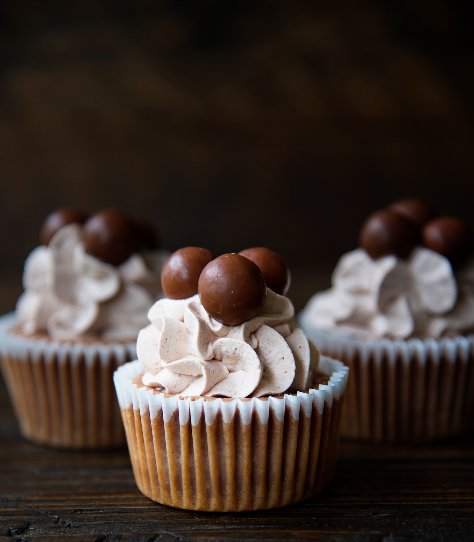 Side view of three Malt Cup Cheesecake Bites with focus on the first cheesecake bite with whipped cream and three malted milk balls. 