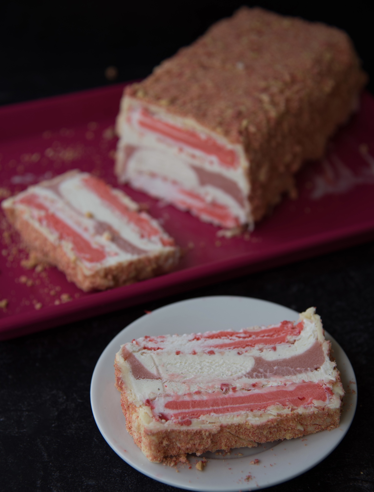 Slice of Strawberry Shortcake Ice Cream Terrine on a small white plate with the remainder of the terrine on a pink sheet pan behind the plate. 