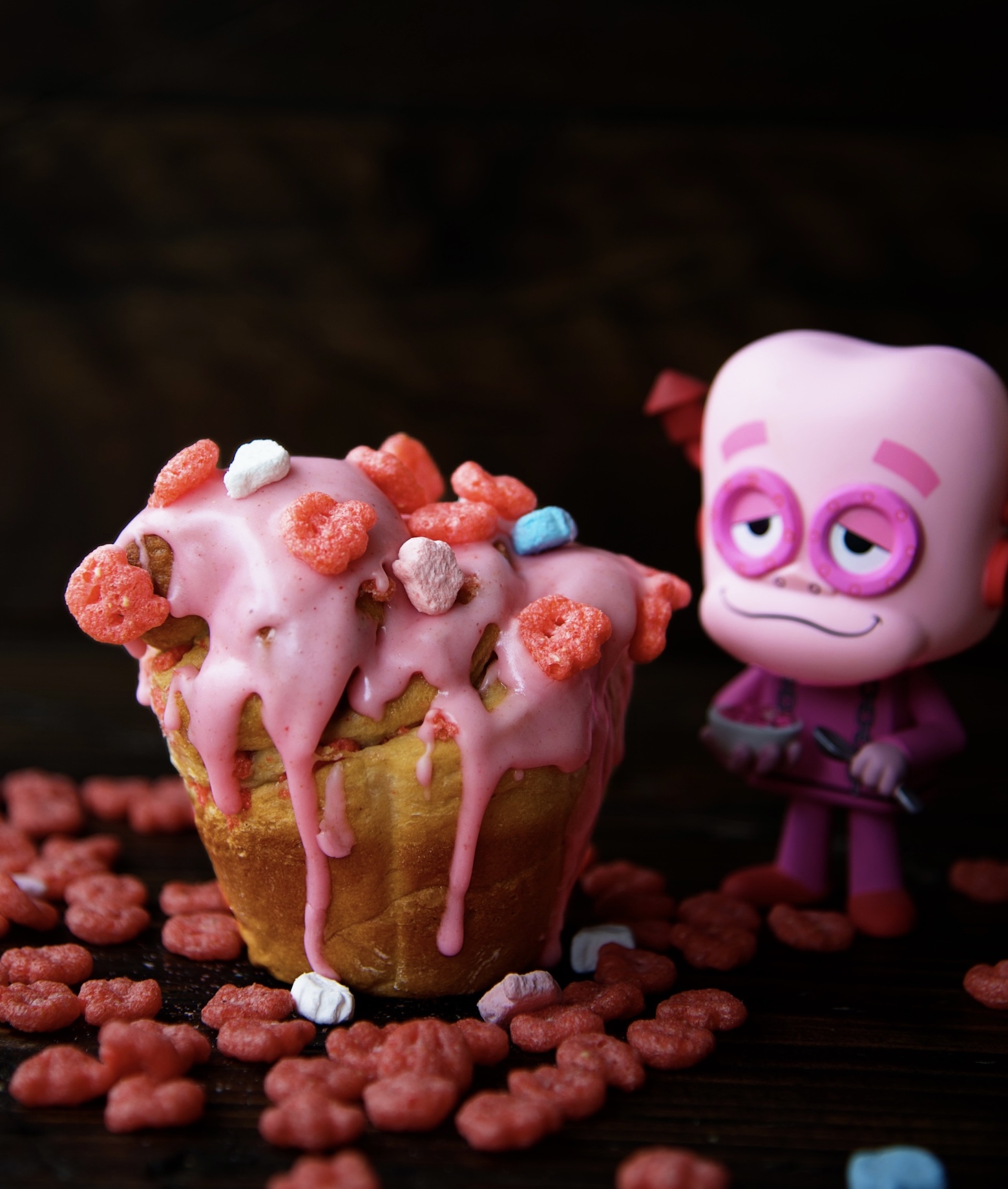 Single Franken Berry Breakfast Bun with pink glaze running down bun with Franken Berry figurine in background. 