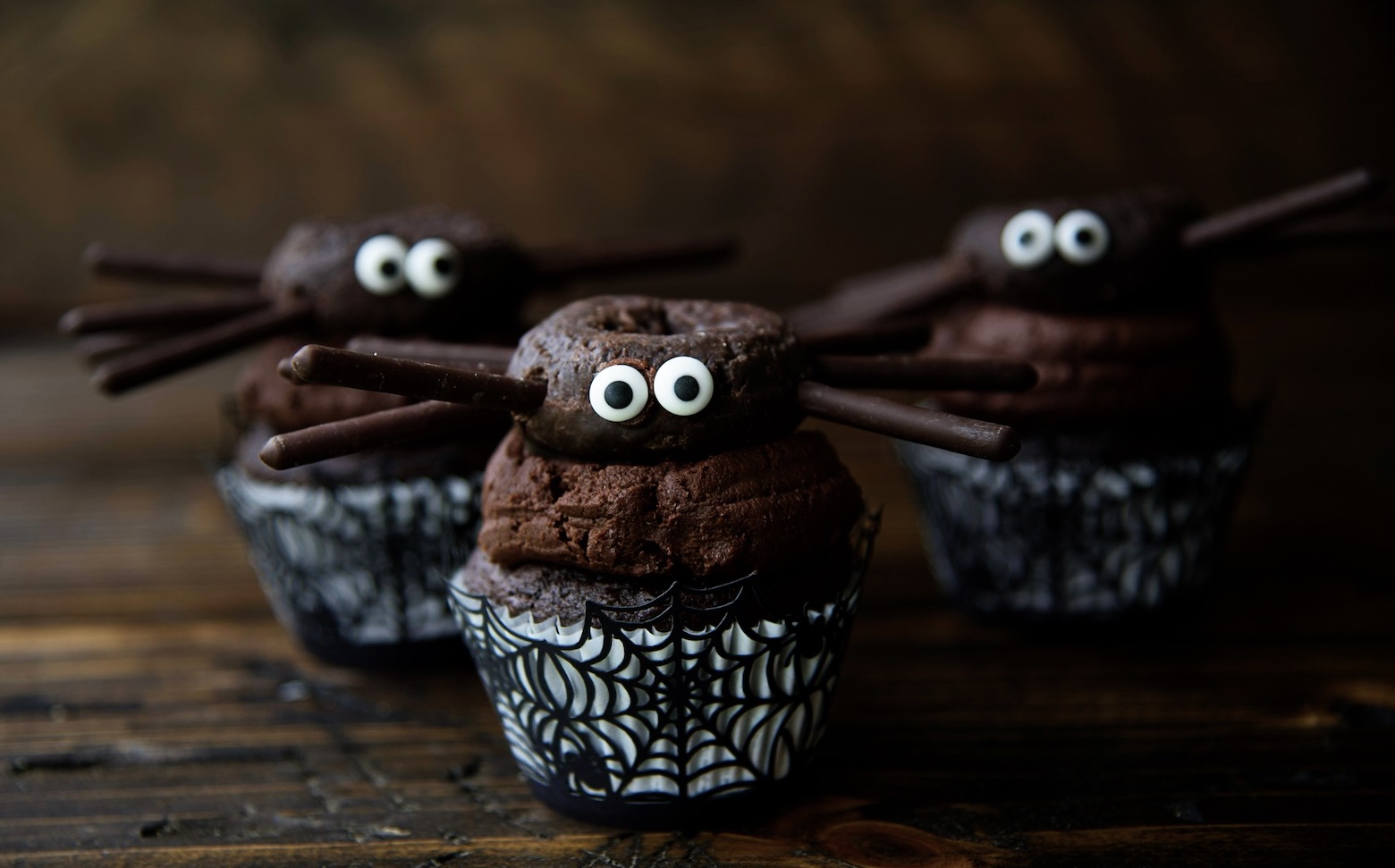 Three chocolate cupcakes with chocolate frosting with chocolate mini donut sitting on top of the icing with eyes and legs to look like spider. 