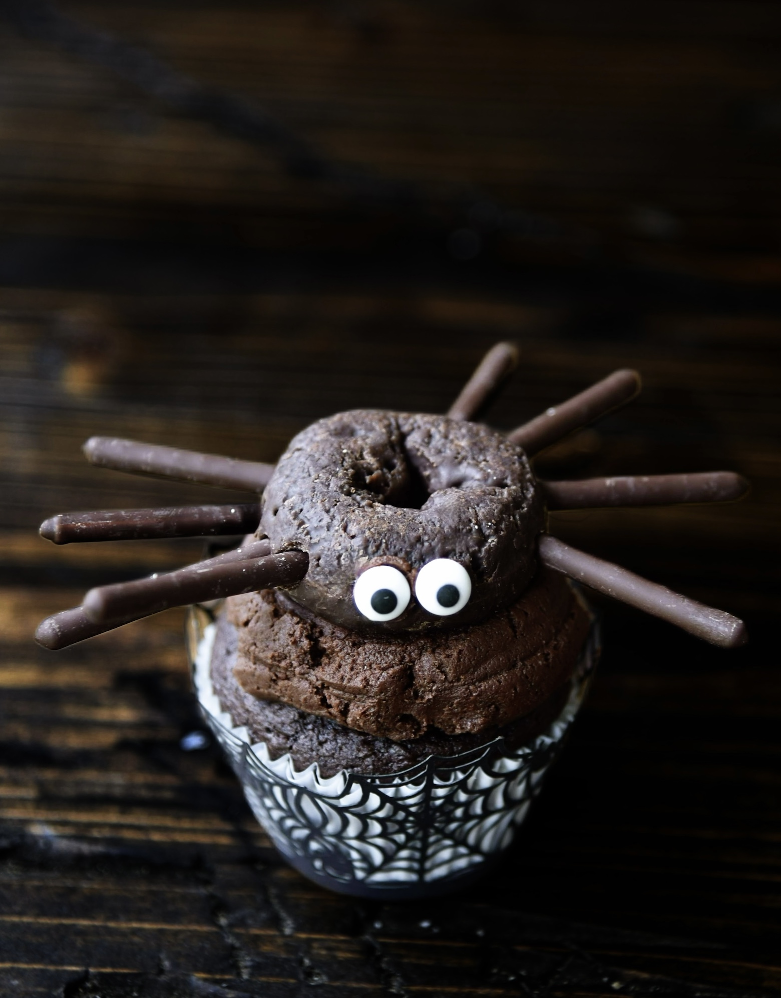 Single chocolate cupcake with chocolate frosting with a mini chocolate donut on top which is made to look like a spider. 