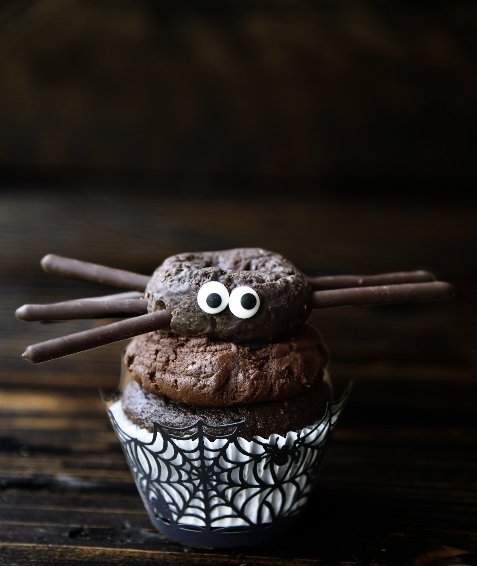 Single chocolate cupcake with chocolate frosting with a mini chocolate donut on top which is made to look like a spider. 