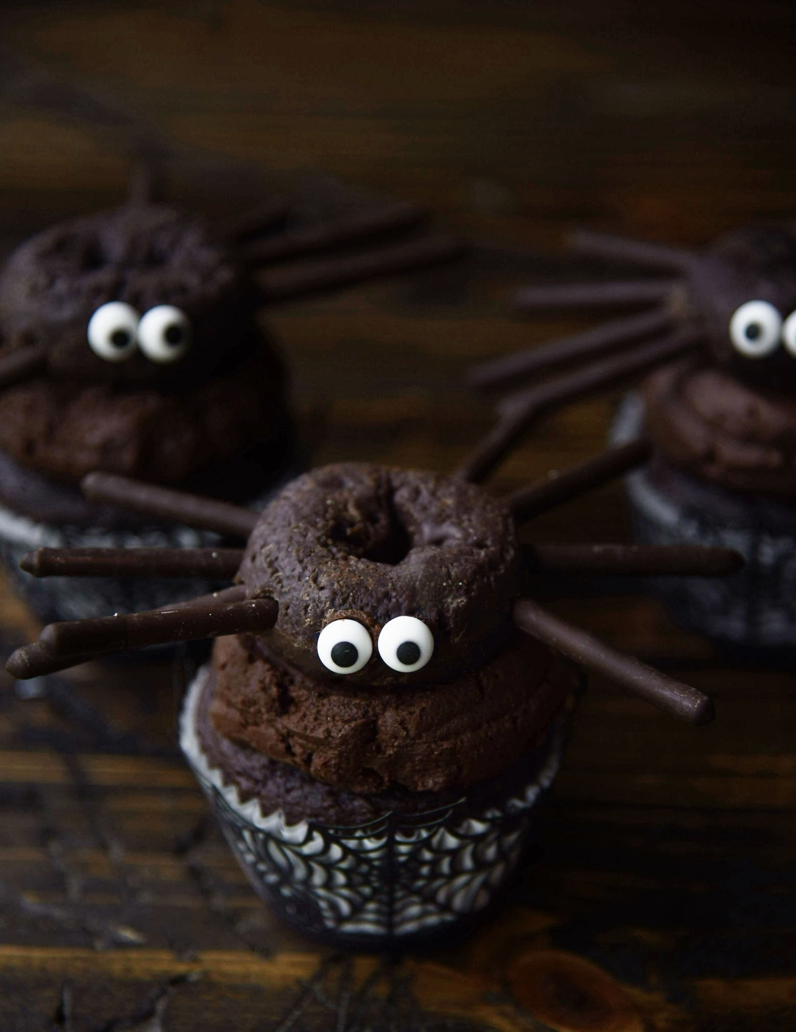 Three chocolate cupcakes with chocolate frosting with chocolate mini donut sitting on top of the icing with eyes and legs to look like spider. 