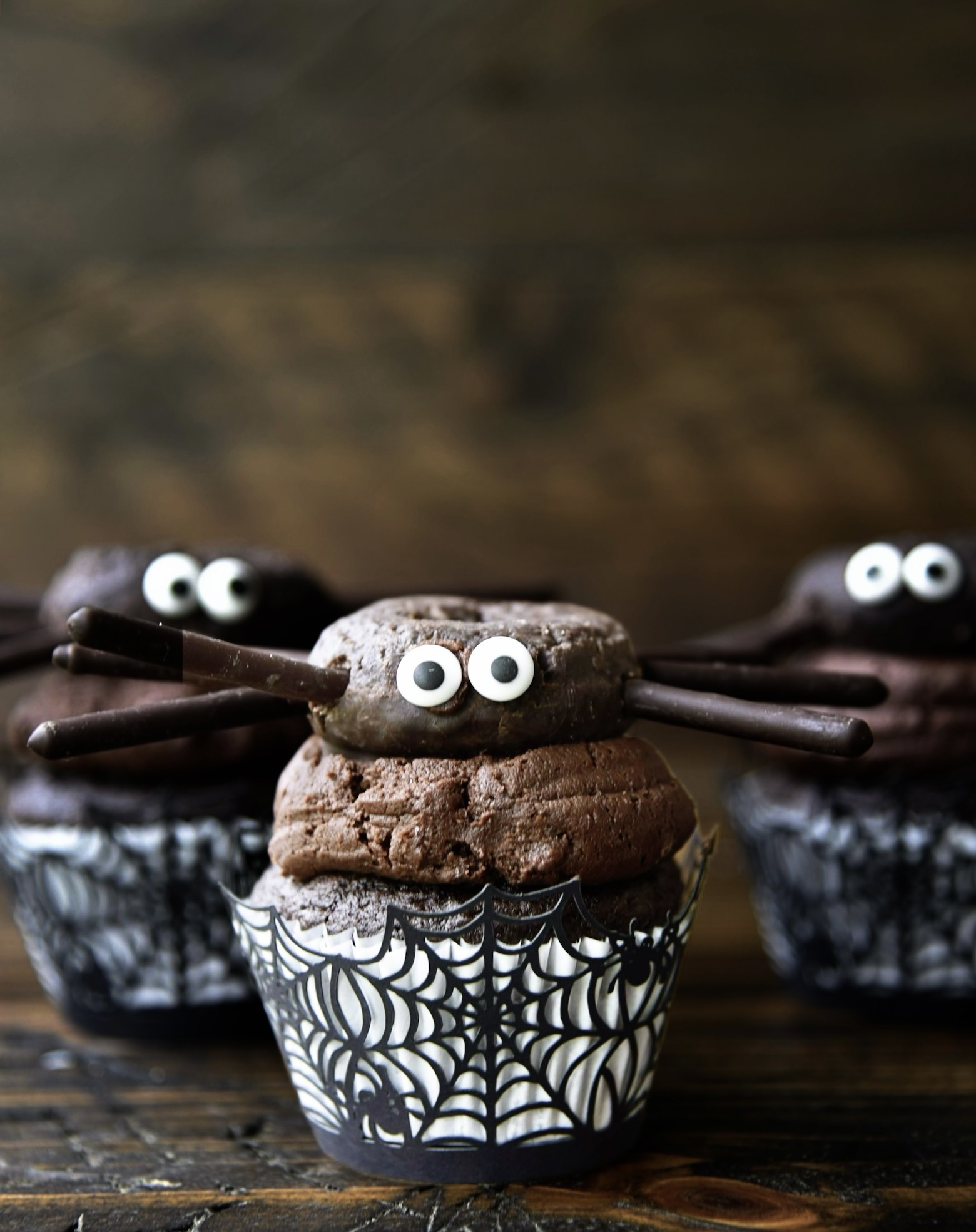 Three chocolate cupcakes with chocolate frosting with chocolate mini donut sitting on top of the icing with eyes and legs to look like spider. Middle chocolate cupcake is in front of the other two cupcakes. 