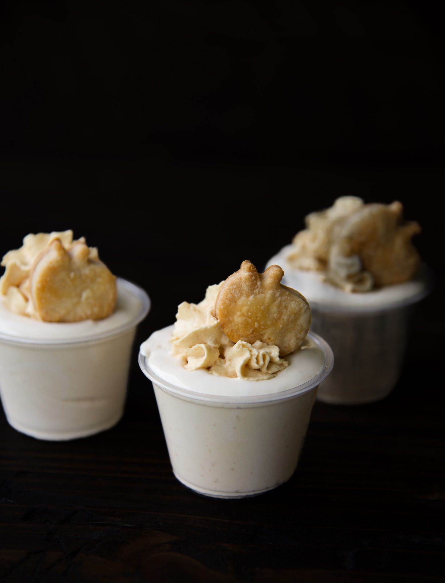 Side view of three apple pie pudding shots in cups with whipped cream and a apple shaped pie crust piece on top.