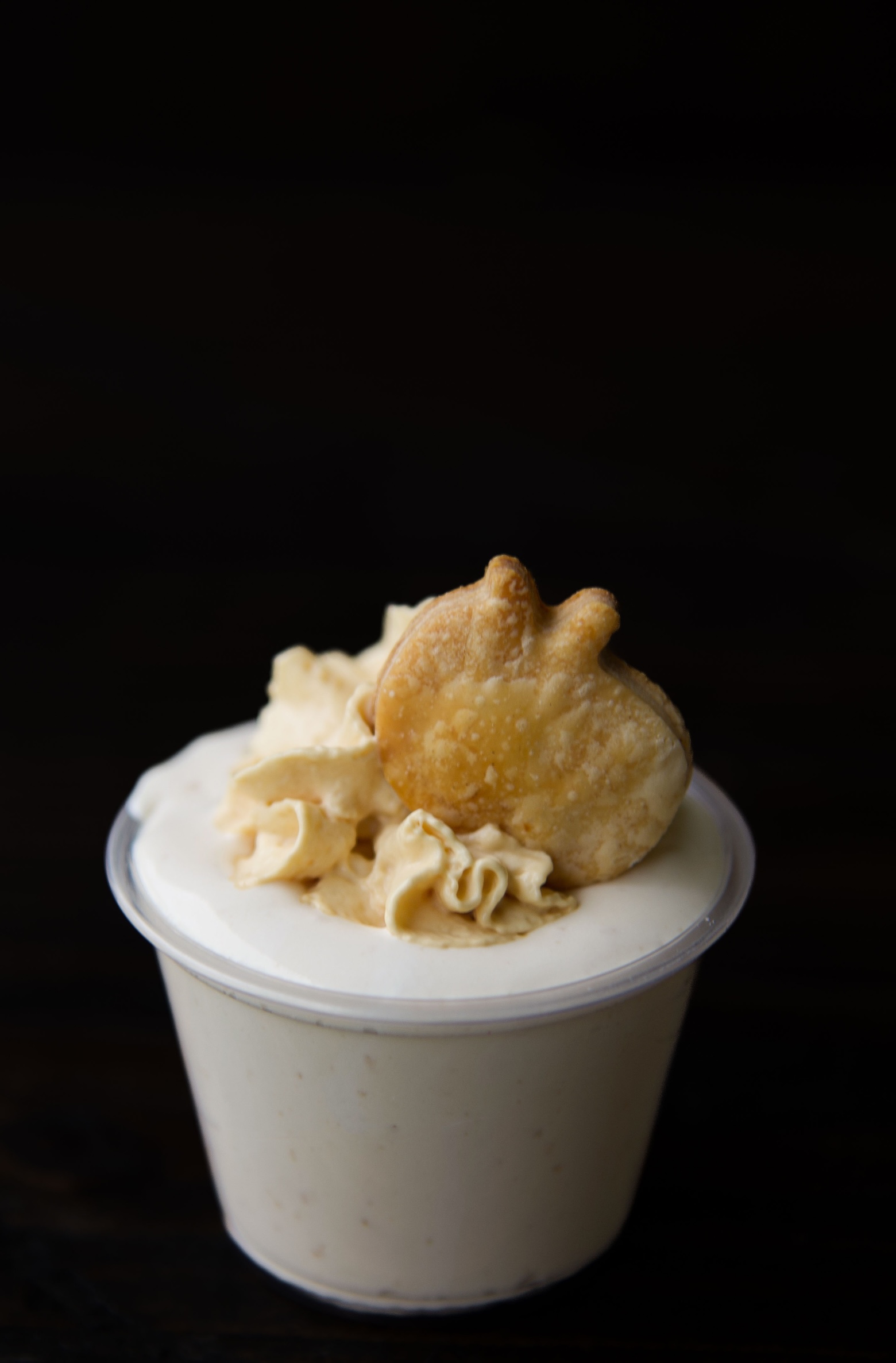 Single apple pie pudding shot in a cup with whipped cream and a apple shaped pie crust piece on top.