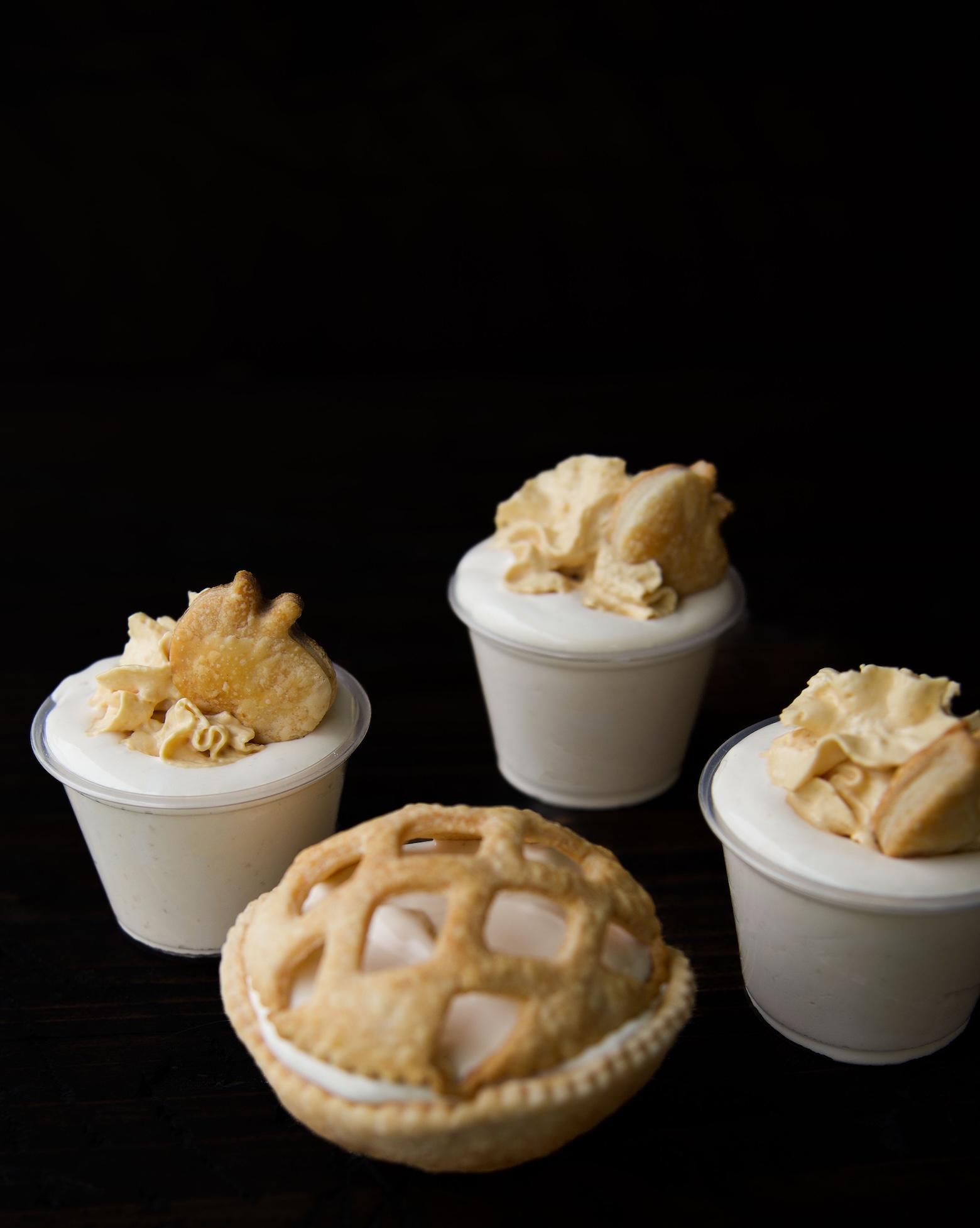 Three apple pie pudding shots in cups with whipped cream and a apple shaped pie crust piece on top with one apple pie pudding shot in front in a pie crust with pie lattice. 