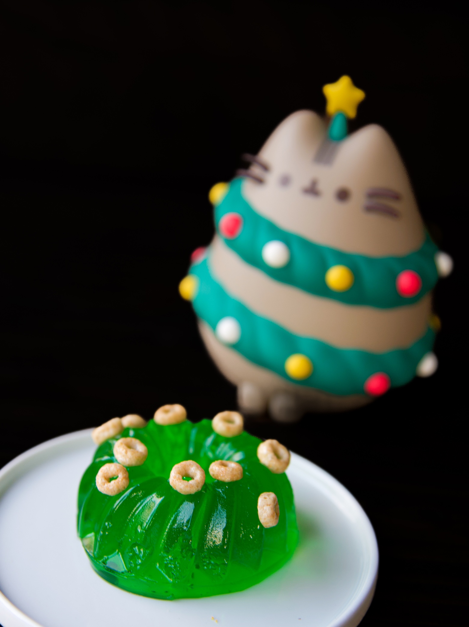 Mini green jello mold with mini cereal scattered on top with cat as a christmas tree to the right of the jello mold.