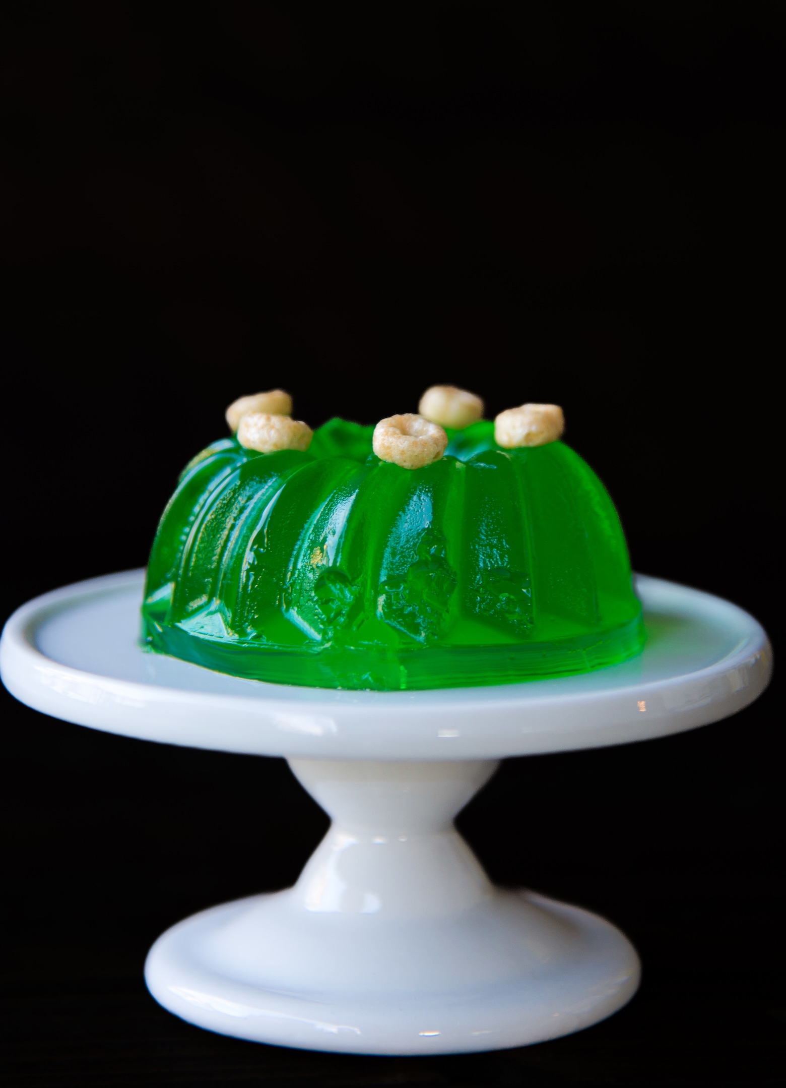 Single mini lime green jello mold on a mini pedestal with scattered mini cereal on top.  