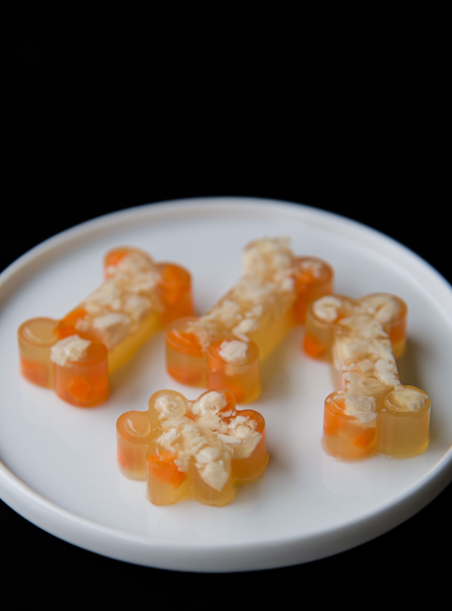 Three chicken jello shot bones and one jello shot paw on a small white plate. You can see the chopped up chicken and carrots. 